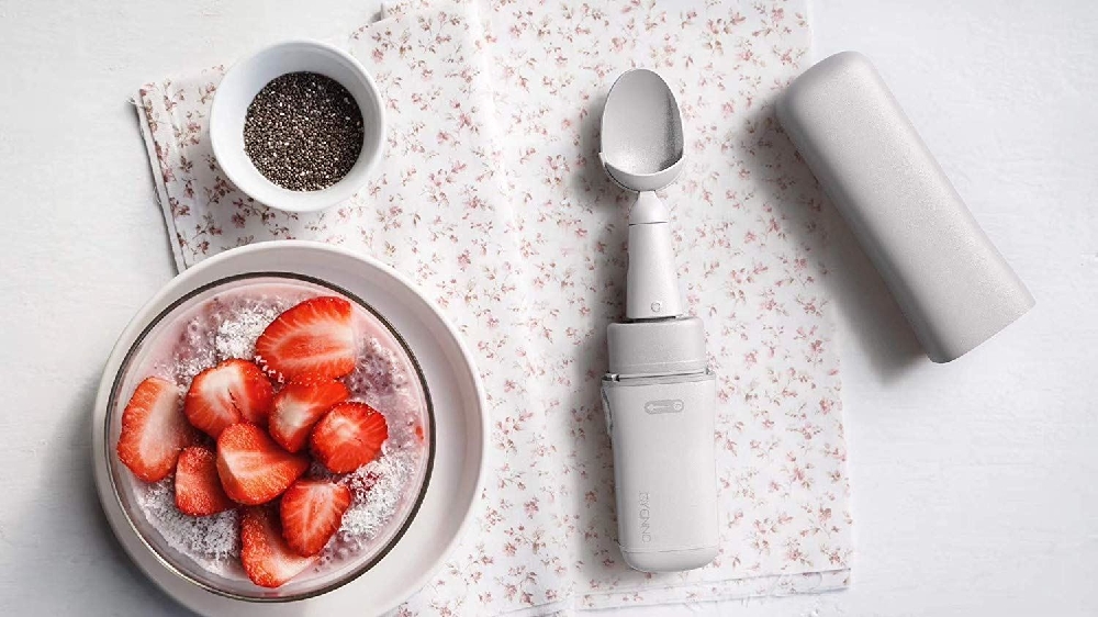 Gyenno smart spoon on a floral napkin beside a bowl of sliced strawberries, a dish of chia seeds, and a white cylinder